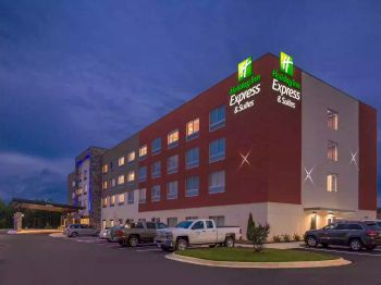 This image shows a Holiday Inn Express and Suites hotel building with a parking lot, several vehicles, and the hotel signage illuminated at night.