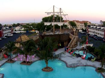 The image shows a pool area with a pirate ship-themed structure, surrounded by palm trees, lounge chairs, and buildings in the background.