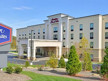 The image shows the exterior of a Hampton Inn & Suites hotel by Hilton with green landscaping and a prominent sign in the foreground.