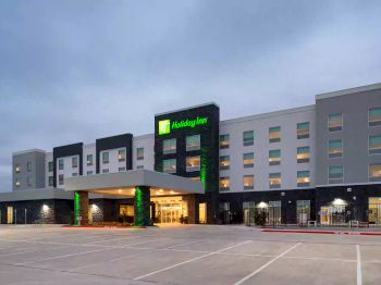 The image shows the exterior of a Holiday Inn hotel with a large parking area in the foreground. The building has several windows and a welcoming entrance.