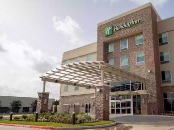 The image shows the front of a Holiday Inn hotel, featuring a modern exterior with a covered entrance, large windows, and landscaped surroundings.