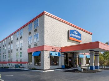 The image shows a hotel building with a sign, blue awnings, a parking area in front, and clear skies in the background.