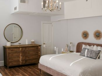 A cozy bedroom featuring a bed with white bedding, a wooden dresser with a round mirror, two wall decorations, and a chandelier hanging from the ceiling.