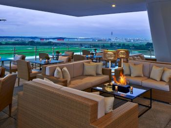 The image shows a modern outdoor lounge area with wicker furniture, cushions, and a firepit, overlooking a lush landscape with distant buildings.