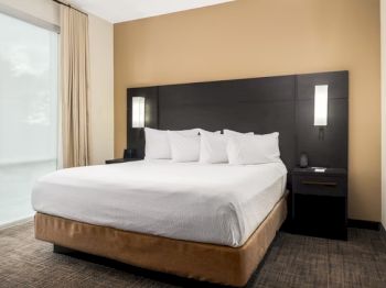 The image shows a neatly made bed with white linens and four pillows in a modern hotel room. There are two nightstands, each with a lamp.