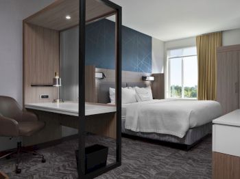 A modern hotel room with a bed, desk area, and chair. It has a large window, a TV, and contemporary decor with neutral tones and geometric patterns.