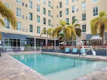 The image shows a swimming pool area with palm trees, lounge chairs, and a multi-story building in the background.