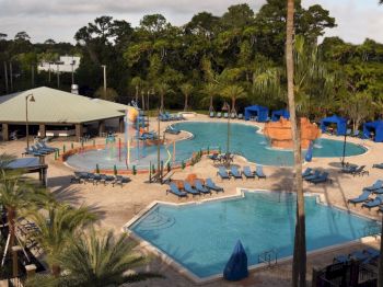 This image features an outdoor resort pool area, complete with lounge chairs, cabanas, a water play area, and surrounded by lush greenery and palm trees.