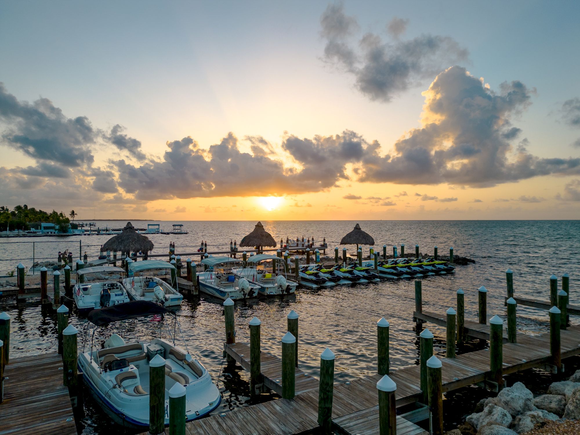 The image shows a serene marina at sunset with boats docked along piers and thatched-roof structures over the water.