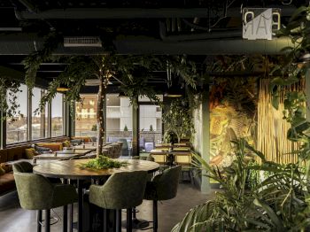 A cozy, modern restaurant with lush greenery, wooden decor, large windows, and comfortable seating arrangements, creating a serene dining atmosphere.